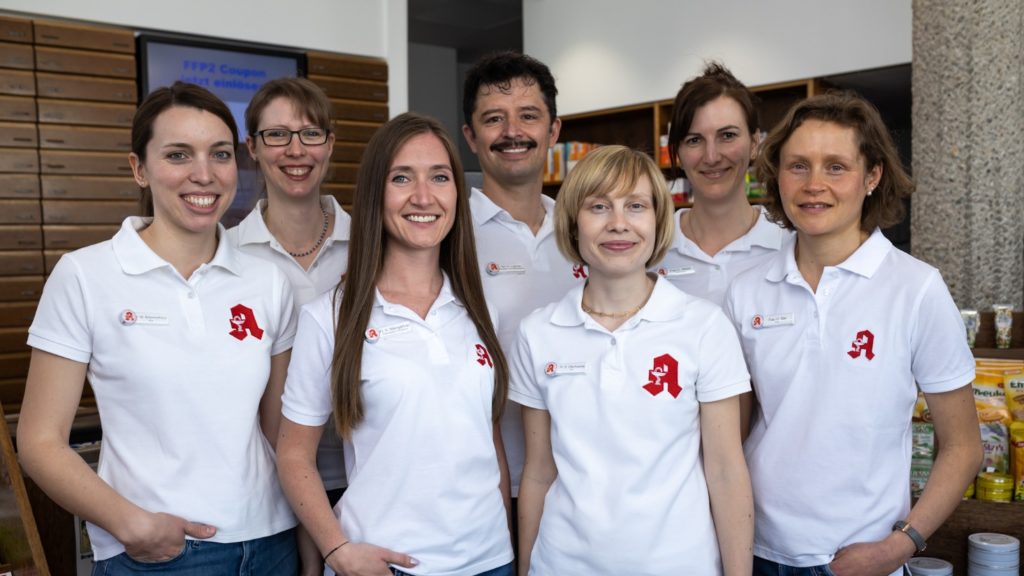 Sieben Personen in passenden weißen Poloshirts mit einem roten Logo stehen lächelnd in einer Reihe. Sie befinden sich im Innenbereich, im Hintergrund sind Regale und eine Theke zu sehen.