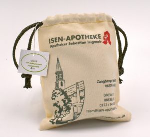 Auf einem kleinen Beutel mit Kordelzug steht „ISEN-APOTHEKE Apotheker Sebastian Parnitzke“ mit einer grünen Skizze eines Gebäudes und Bäumen. Auf einem Etikett steht „OFFRO 100% Olive Pine White Lana“. Kontaktdaten sind auf dem Beutel aufgedruckt.