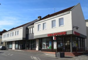 In einem zweistöckigen Gebäude mit weißer Fassade ist eine Apotheke untergebracht. Über dem Eingang steht in roten Lettern „ISEN-APOTHEKE“. Große Fenster im Erdgeschoss zeigen Produkte. Der Himmel ist klar und blau.