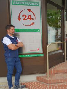 Ein Mann in einer blauen Weste steht vor einer Apotheke neben einem Schild mit der Aufschrift „Abholstation 24h“, was auf eine rund um die Uhr geöffnete Abholstation hinweist. Das Gebäude hat einen Eingang aus Ziegelsteinen und Glastüren. Der Mann blickt mit verschränkten Armen zur Seite.