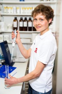 Eine Person mit kurzen Haaren und weißem Poloshirt hält eine Pipette über ein Reagenzglas. Sie steht in einem Labor, im Hintergrund sind Regale voller Flaschen zu sehen, und lächelt in die Kamera.