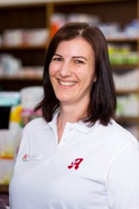 Lächelnde Person mit schulterlangem dunklem Haar und weißem Poloshirt steht im Innenbereich. Im Hintergrund sind Regale mit verschiedenen Produkten verschwommen.