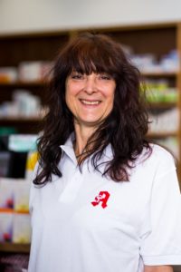 Eine Frau mit langen, dunklen Haaren lächelt und trägt ein weißes Poloshirt mit einem roten Logo. Sie steht vor Regalen voller verschiedener Artikel.
