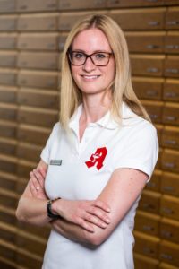 Eine Frau mit langen blonden Haaren und Brille steht lächelnd mit verschränkten Armen da. Sie trägt ein weißes Poloshirt mit einem roten Logo auf der Brust. Im Hintergrund ist eine Wand mit Holzschubladen zu sehen.