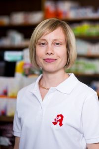Eine Frau mit kurzen blonden Haaren lächelt leicht und trägt ein weißes Poloshirt mit einem roten Logo auf der Brust. Im Hintergrund sind Regale mit verschiedenen bunten Produkten zu sehen.
