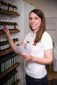 Eine Frau in einer Apotheke lächelt, während sie Medikamentenflaschen in einem Regal ordnet. Sie hält ein Stück Papier in der Hand und trägt ein weißes Poloshirt.
