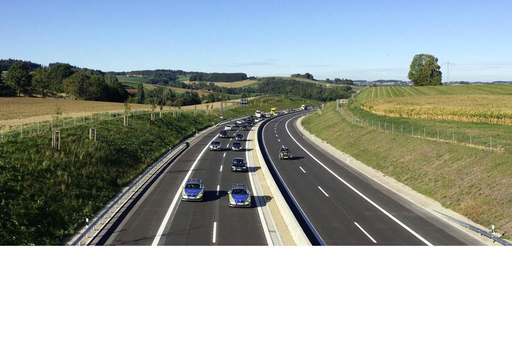 Auf einer Autobahnszene sind mehrere Autos zu sehen, die in beide Richtungen fahren. Die Straße ist von grünen Feldern und Bäumen unter einem klaren blauen Himmel gesäumt. Auf der rechten Seite sind Verkehrsleitkegel und ein Lieferwagen zu sehen, die auf mögliche Straßenarbeiten hinweisen.