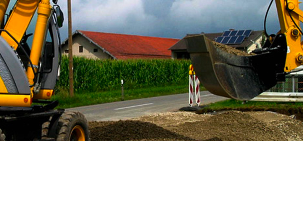 Ein gelber Baggerlader transportiert Kies neben einer Landstraße. Im Hintergrund sind ein Gebäude mit rotem Dach und ein Maisfeld zu sehen. Der Himmel ist bewölkt und auf einem Mast ist ein Solarpanel zu sehen.