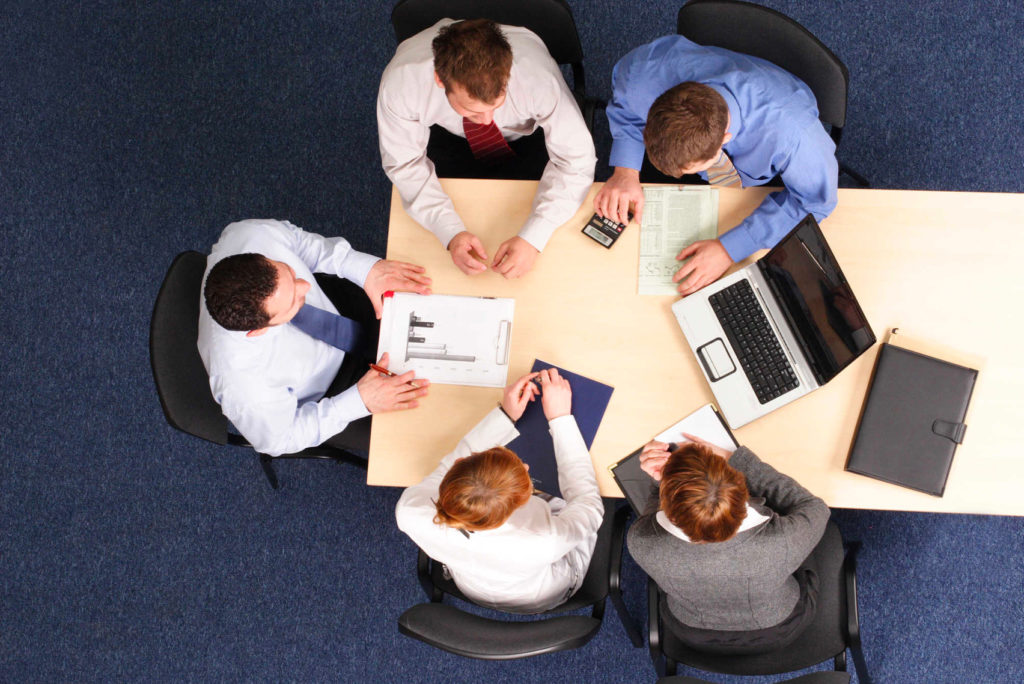 Luftaufnahme von fünf Personen in Geschäftskleidung, die an einem rechteckigen Tisch ein Meeting abhalten. Sie betrachten Dokumente und einen Laptop auf dem Tisch. Die Kulisse ist ein Konferenzraum mit blauem Teppich und schwarzen Stühlen.