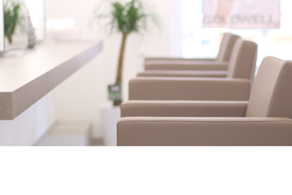 Eine Reihe beiger Friseurstühle gegenüber einem Spiegel und einer Arbeitsplatte in einem hellen, modernen Raum. Im Hintergrund ist eine Pflanze zu sehen, die dem sauberen, minimalistischen Dekor einen Hauch von Grün verleiht.