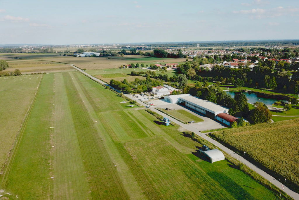 Luftaufnahme einer ländlichen Landschaft mit ausgedehnten grünen Feldern. Eine kleine Landebahn ist sichtbar, in deren Nähe sich mehrere Hangars und Gebäude befinden. Zwischen Bäumen liegt ein Teich, und in der Ferne ist unter einem klaren blauen Himmel ein Wohngebiet zu sehen.