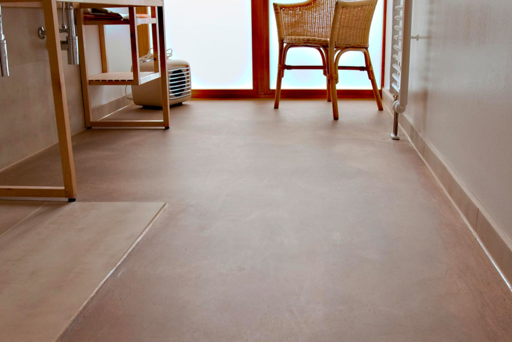 Ein minimalistischer Raum verfügt über einen glatten beigen Boden und zwei Rattanstühle neben einem Milchglasfenster. Auf der linken Seite befinden sich ein kleiner weißer Heizkörper und ein Holzregal. Die Gesamteinrichtung ist schlicht und in hellen Tönen gehalten.