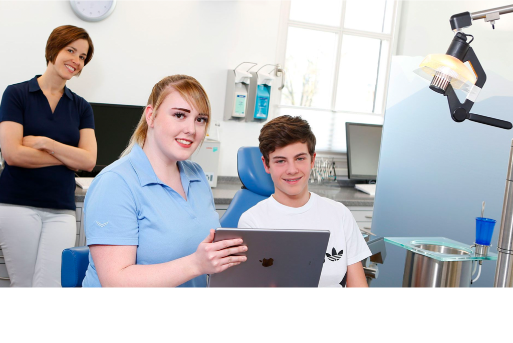 Eine Szene in einer Zahnarztpraxis mit einem jungen Patienten, der auf einem Stuhl sitzt. Ein Zahnarzt neben ihm hält ein Tablet in der Hand. Eine weitere Person steht lächelnd im Hintergrund. Der Raum ist mit medizinischen Geräten ausgestattet und hell beleuchtet.