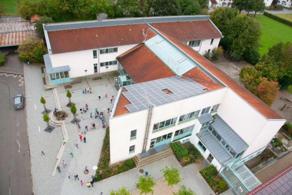 Luftaufnahme eines modernen Gebäudes mit weißer Außenfassade und rotbraunen Schrägdächern, einige mit Solarmodulen. Ein Innenhof mit Bäumen und ein paar Menschen ist zu sehen, umgeben von Gras und anderen Gebäuden.