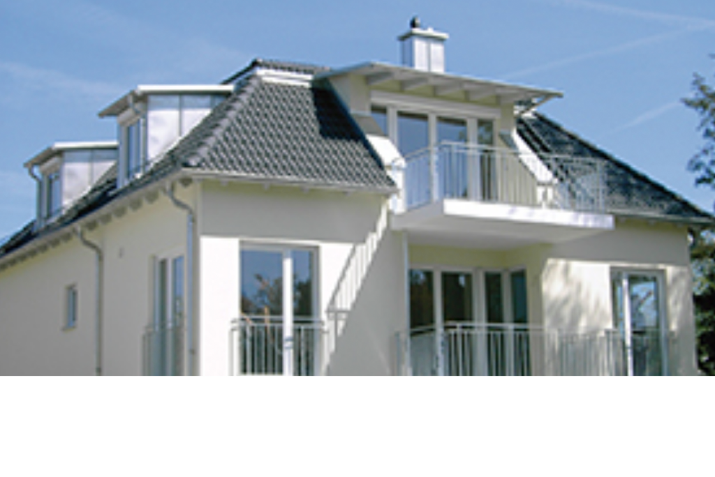 Ein modernes zweistöckiges Haus mit einem dunkelgrauen Schrägdach und weißen Wänden. Im Obergeschoss sind große Fenster und ein Balkon zu sehen. Das Haus ist von Bäumen umgeben und der Himmel ist klar und blau.