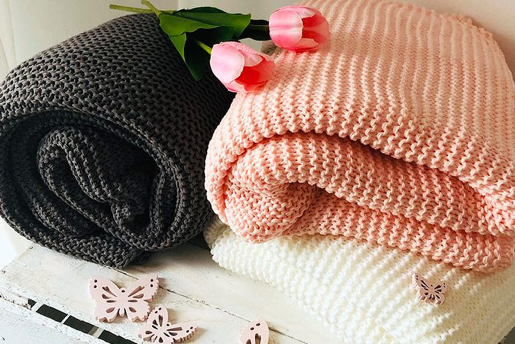 Zwei gestrickte Decken, eine dunkelgrau und eine hellrosa, sind zusammengefaltet und mit einer cremefarbenen Decke auf einer weißen Holzoberfläche gestapelt. Daneben liegen rosa Tulpen und kleine dekorative Schmetterlinge.