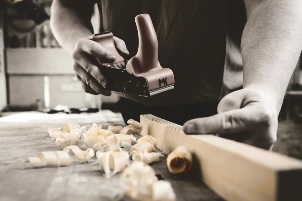 Eine Person glättet mit einem Handhobel ein Holzbrett, umgeben von Holzspänen auf einer Werkbank. Das Bild hat einen Sepiaton, der die Handwerkskunst und den Fokus auf die Holzbearbeitung betont.