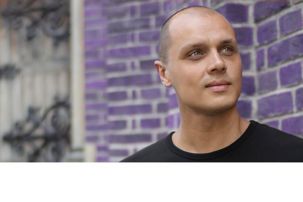 Ein Mann mit rasiertem Kopf und schwarzem T-Shirt steht vor einem Hintergrund aus violetten Ziegeln und blickt nachdenklich zur Seite. Im unscharfen Hintergrund ist teilweise eine dekorative Metallstruktur zu erkennen.