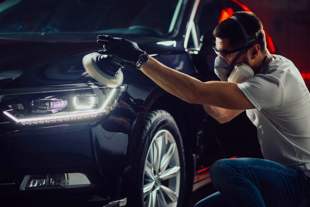 Eine Person mit weißem Hemd, Handschuhen und Schutzmaske poliert in einer schwach beleuchteten Garage mit einer Poliermaschine den Scheinwerfer eines dunklen Autos. Das Auto ist glänzend und glatt und reflektiert das Licht.