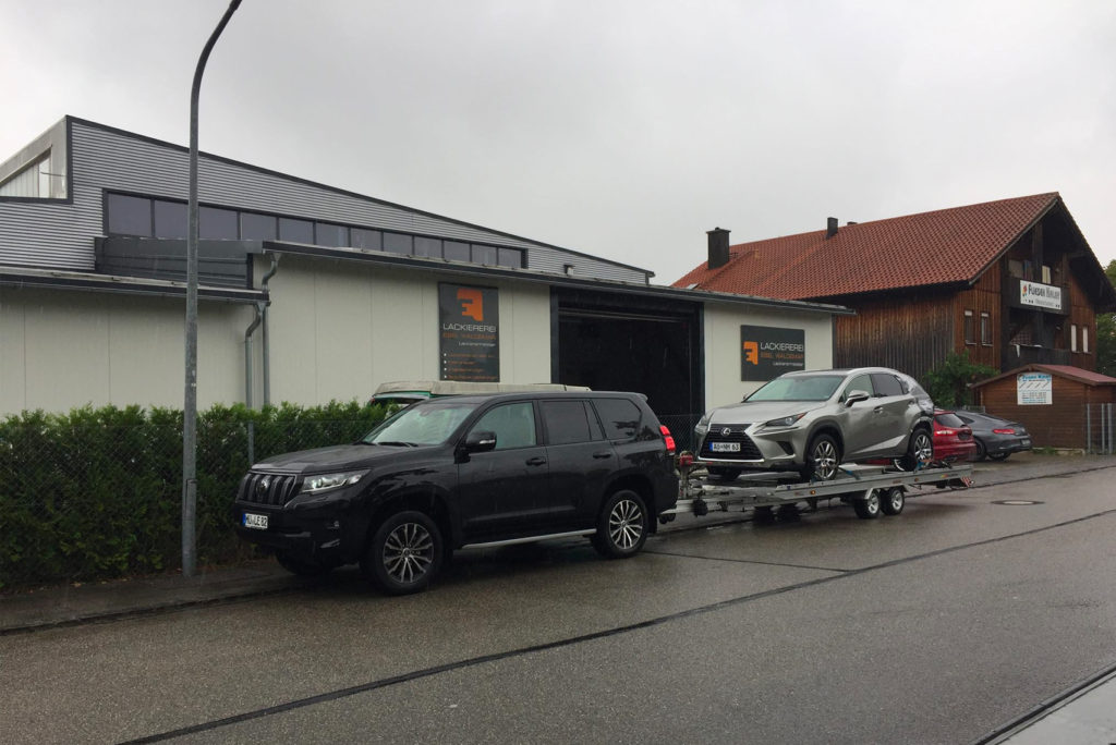 Ein schwarzer SUV zieht ein silbernes Auto auf einem Anhänger vor einem Gebäude mit Schrägdach. Die Straße ist nass, was auf kürzlichen Regen hindeutet. Im Hintergrund sind eine Hecke und ein weiteres Gebäude mit einem roten Ziegeldach zu sehen.