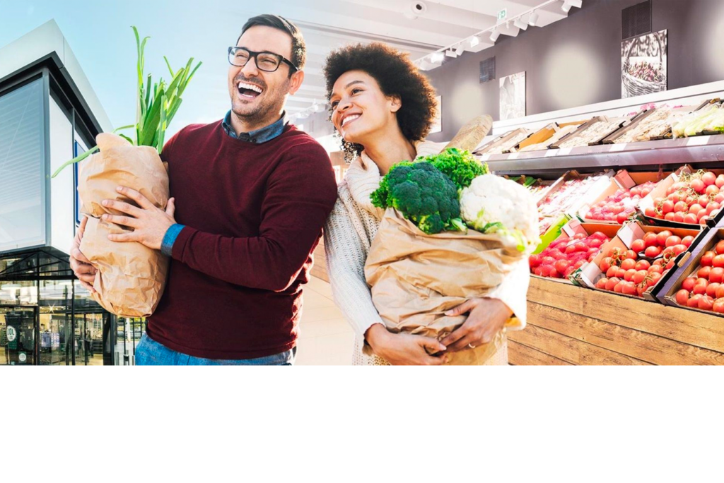 Ein Mann und eine Frau tragen fröhlich Einkaufstüten mit frischen Produkten wie Brokkoli und Frühlingszwiebeln durch einen hellen Supermarkt. Hinter ihnen stehen Holzkisten mit Tomaten und anderem Gemüse.