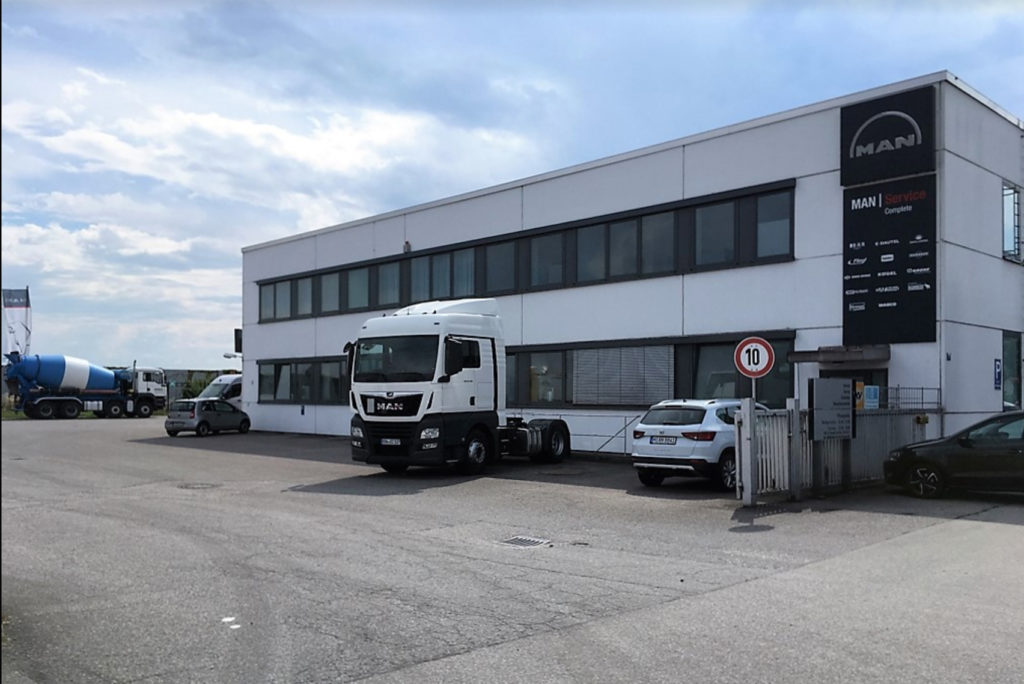 Außenansicht eines MAN-Servicecenters mit einem großen weißen LKW, der davor parkt. Das Gebäude hat Glasfenster und ein Schild mit dem MAN-Logo. Im Hintergrund ist ein Betonmischer zu sehen, und in der Nähe ist ein Schild mit einer Geschwindigkeitsbegrenzung von 10 km/h angebracht.