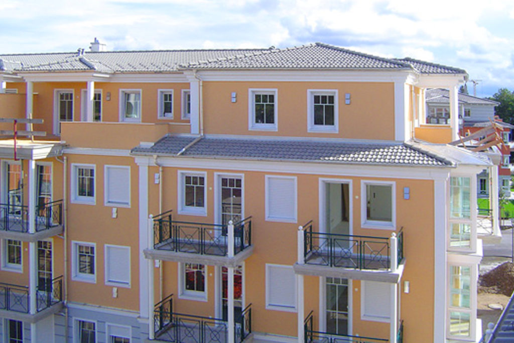 Ein modernes, mehrstöckiges Wohnhaus mit orangefarbenen Wänden und weißen Zierleisten. Es verfügt über Balkone mit Geländern, zahlreiche Fenster und ein graues Ziegeldach. In der Umgebung sind Bäume und andere Wohngebäude unter einem teilweise bewölkten Himmel zu sehen.