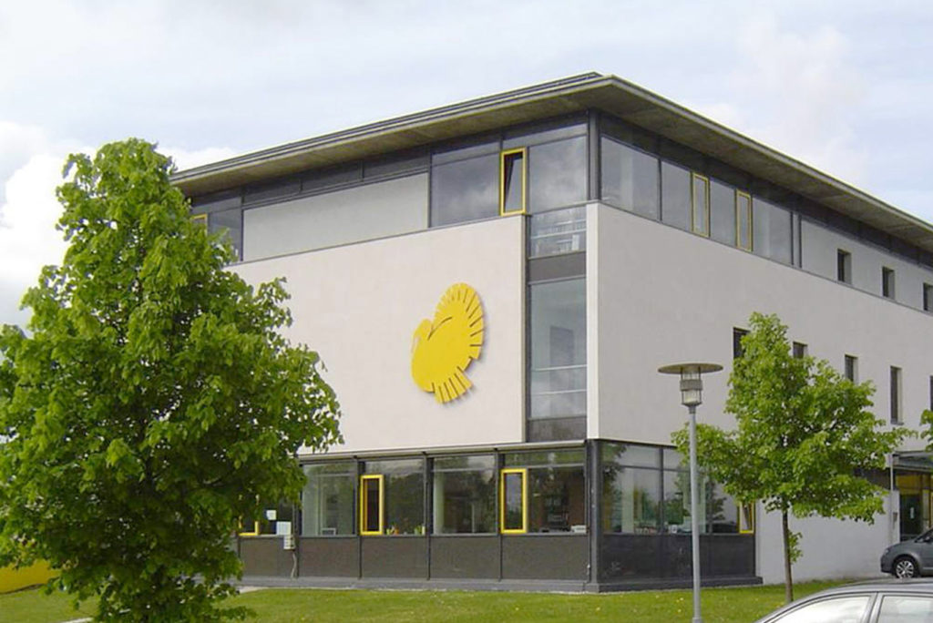 Modernes Gebäude mit einem großen gelben Vogellogo auf der Fassade und Reihen von Fenstern mit gelben Rahmen. Das Gebäude ist von grünen Bäumen und einer Straßenlaterne umgeben, der Himmel ist teilweise bewölkt.