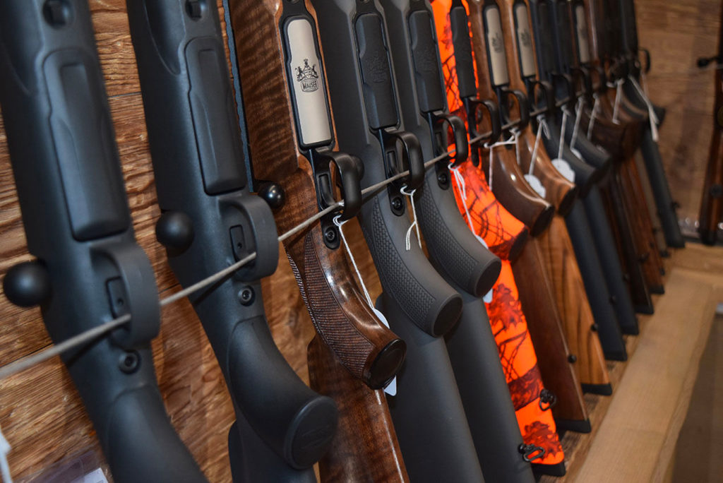 An einer Holzwand hängt vertikal eine Reihe Gewehre. Manche sind dunkel lackiert, andere haben leuchtend orangefarbene Tarnmuster. Die Gewehre sind mit einem dünnen Draht befestigt.