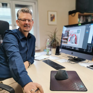 Ein lächelnder Mann mit Brille sitzt an einem Schreibtisch und greift nach einem Stift. Auf einem großen Computerbildschirm wird ein Grafikdesignprojekt angezeigt, möglicherweise die Arbeit an einem Amfingergutschein-Layout. Auf dem Schreibtisch stehen eine Tastatur, eine Maus und eine Wasserflasche. Im Hintergrund hängen eine Pflanze und ein gerahmtes Bild an der Wand.