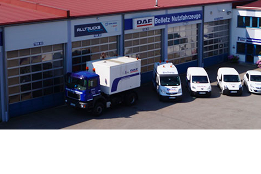 Vor einem Gebäude mit rotem Dach parken eine Reihe Nutzfahrzeuge, darunter ein großer Lastwagen und mehrere Lieferwagen. Auf dem Gebäude sind Schilder mit den Namen „DAF“ und „Belletz Nutzfahrzeuge“ zu sehen.