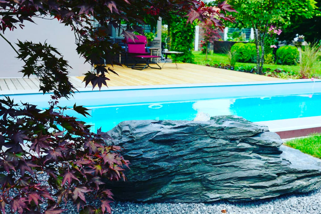 Ein ruhiger Hinterhof verfügt über einen türkisfarbenen Pool, umgeben von üppigem Grün. Im Vordergrund liegen ein großer Schieferfelsen und Kies. Im Hintergrund ist eine Holzterrasse mit einem roten Liegestuhl zu sehen, die teilweise von belaubten Zweigen beschattet wird.