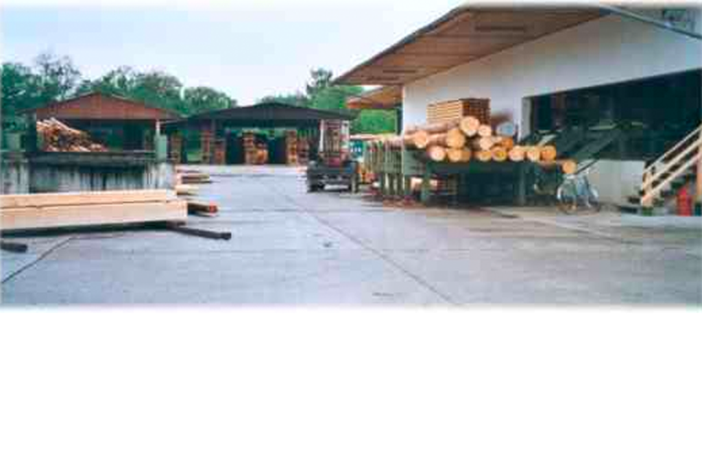 Sägewerkhof mit mehreren Stapeln geschnittener Stämme und Schnittholz. In der Mitte parkt ein Lastwagen, und um den Bereich herum stehen mehrere Gebäude mit Holzstapeln. Im Hintergrund bedeckter Himmel.