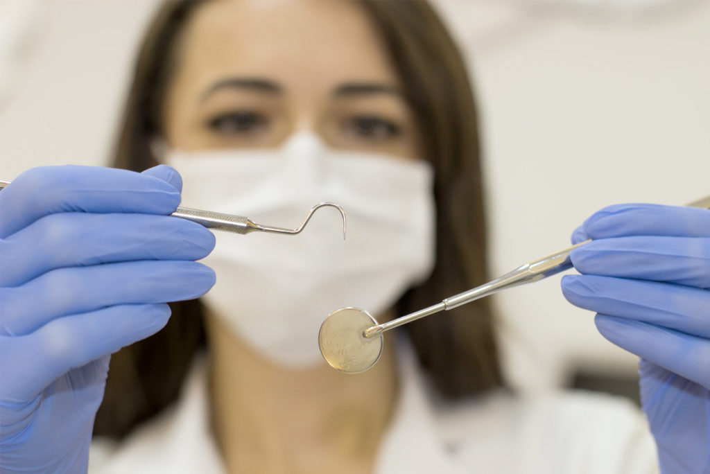 Ein Zahnarzt mit weißer Maske und blauen Handschuhen hält eine Zahnsonde und einen Spiegel in die Kamera. Der Fokus liegt auf den Instrumenten, der Zahnarzt ist im Hintergrund leicht unscharf.
