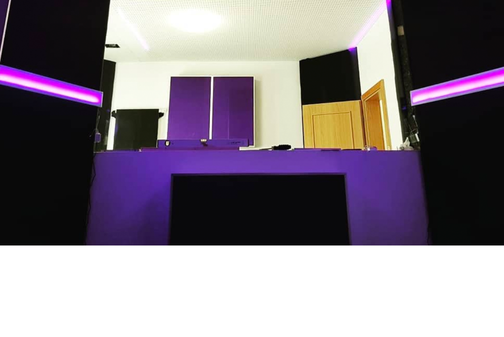 Ein Raum mit violett beleuchteten Akustikplatten an den Wänden, einer weißen Decke und zwei Holztüren. In der Mitte stehen rechteckige Schränke und ein Schreibtisch, beleuchtet von LED-Deckenleuchten. Die Atmosphäre ist modern und minimalistisch.