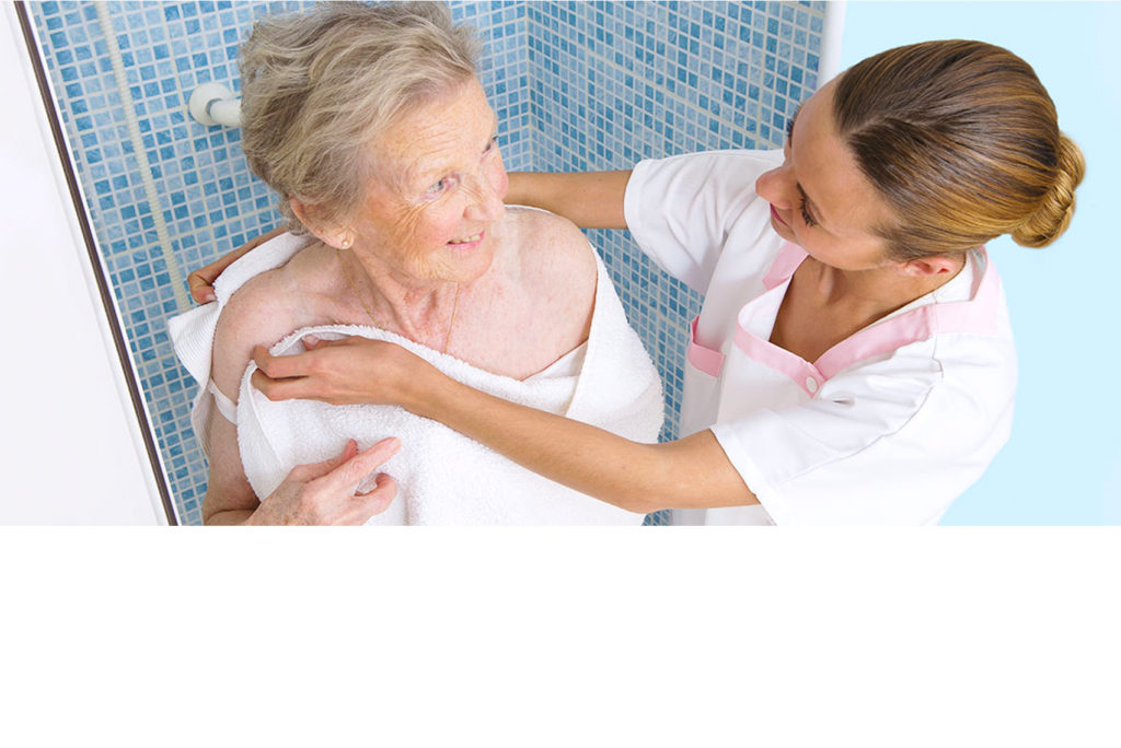 Eine Pflegekraft hilft einer älteren Frau, die in ein Handtuch gehüllt ist, im Badezimmer. Die Pflegekraft lächelt und streckt sanft die Hand nach der Schulter der Frau aus, während die Frau sie mit warmem Gesichtsausdruck ansieht. Die Umgebung ist mit blau gefliesten Wänden ausgestattet.