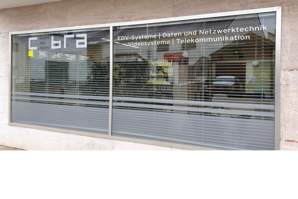 Eine Schaufensterfront mit großen Fenstern und Jalousien, die das Straßenleben widerspiegeln. Der Text auf dem Fenster beschreibt in deutscher Sprache IT-Systeme, Daten- und Netzwerktechnologie, Videosysteme und Telekommunikation.