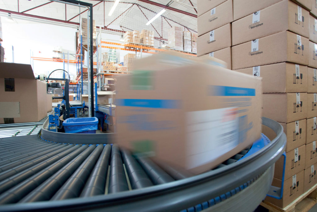 Ein großer, unscharfer Karton bewegt sich schnell auf einem Förderband in einem Lagerhaus. Rechts sind Stapel von Kartons aufgetürmt, im Hintergrund sind Regale und weitere Kartons zu sehen. Die Szene lässt auf einen geschäftigen Logistikbetrieb schließen.