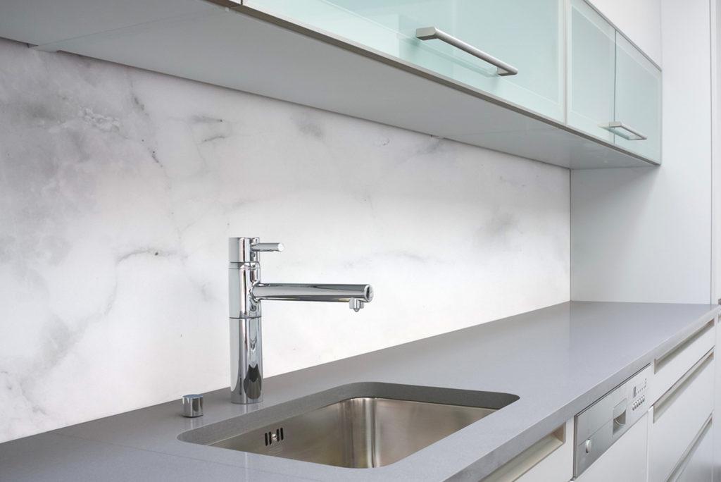 Modernes Kücheninterieur mit einer eleganten grauen Arbeitsplatte mit integrierter Edelstahlspüle und minimalistischem Chromhahn. Die Küchenrückwand ist in glattem, weißem Marmor gehalten und die Oberschränke haben Milchglastüren für einen klaren, zeitgenössischen Look.