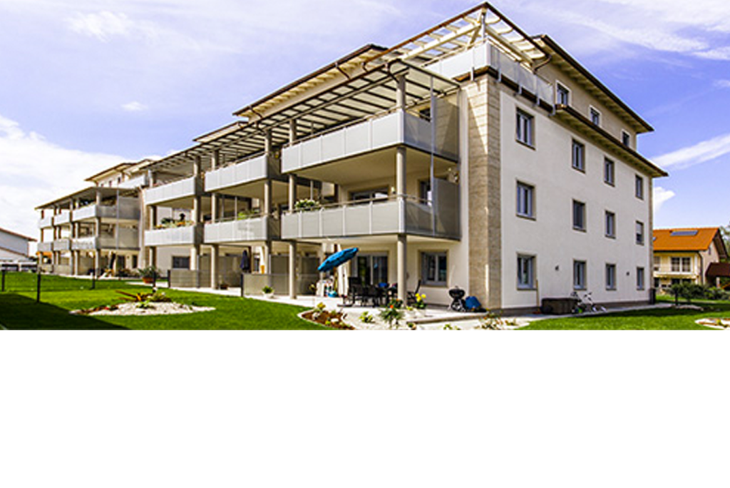 Ein modernes, mehrstöckiges Apartmentgebäude mit Balkonen und Flachdach vor einem blauen Himmel. Es verfügt über einen gepflegten Rasen mit einigen Landschaftsgestaltungen, darunter ein kleiner Garten und Wege rund um das Gebäude.