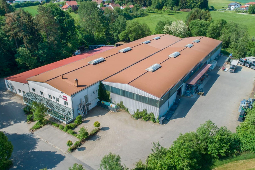 Luftaufnahme eines großen Industrielagers mit rostfarbenem Dach, umgeben von grünen Bäumen und Gras. Das Gebäude hat mehrere Oberlichter und einen großen gepflasterten Bereich mit geparkten Fahrzeugen in der Nähe.