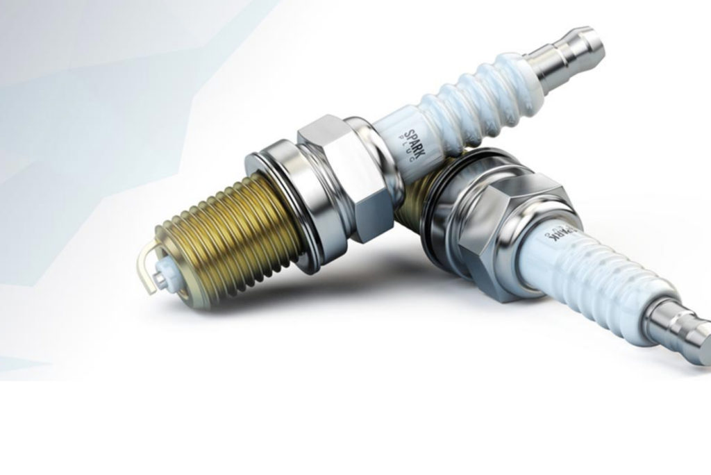 Zwei metallische Zündkerzen mit weißen Keramikisolatoren liegen auf einer hellgrauen Oberfläche. Die linke Zündkerze ist horizontal angeordnet, während die rechte leicht angewinkelt ist. Der Hintergrund ist ein Farbverlauf von Hellgrau nach Weiß.