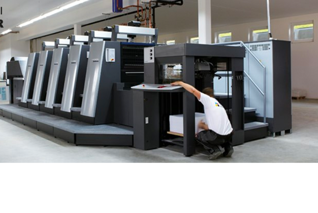 Person, die in einer gut beleuchteten Halle an einer großen industriellen Druckmaschine arbeitet. Sie trägt ein weißes Hemd und schwarze Shorts. Die Maschine besteht aus mehreren Abschnitten mit Bedienfeldern und Papierstapeln.