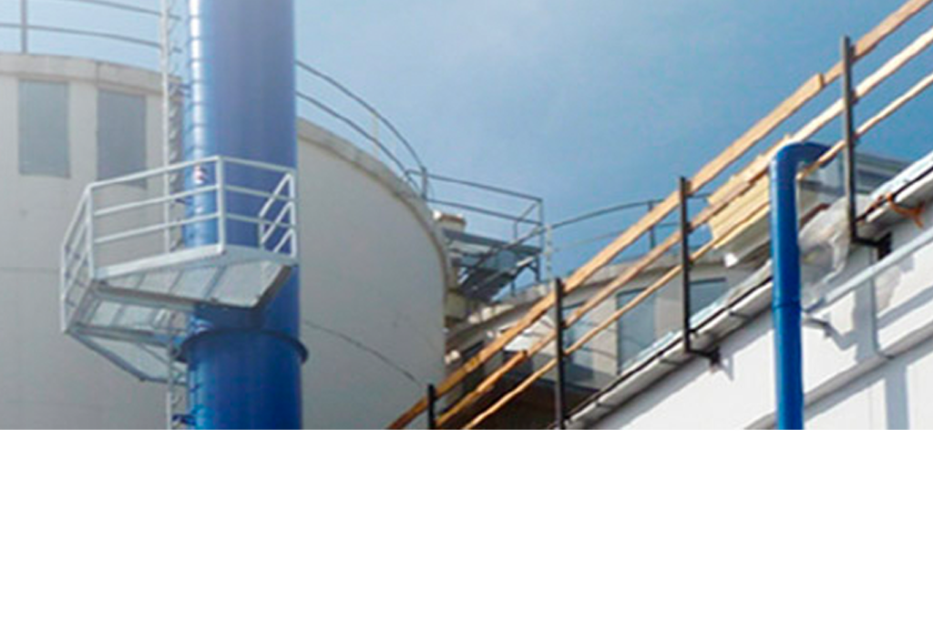 Industrielle Szene mit einer großen weißen zylindrischen Struktur, möglicherweise einem Lagertank, mit blauen vertikalen Rohren und Geländern. Im Hintergrund ist ein klarer, sonniger Himmel zu sehen.