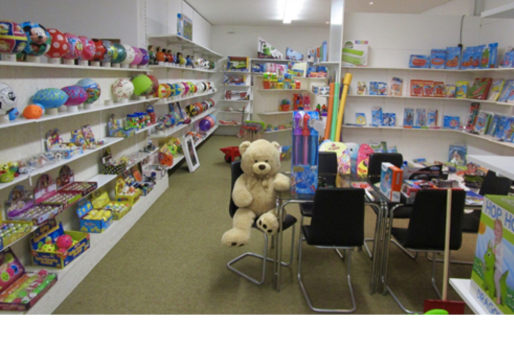 Ein Spielzeugladen mit Regalen voller bunter Spielsachen, darunter Bälle, Schachteln und Puppen. In der Mitte stehen schwarze Stühle um einen Tisch herum, auf einem Stuhl sitzt ein großer Teddybär. Überall im Raum sind verschiedene Spiele und Puzzles ausgestellt.