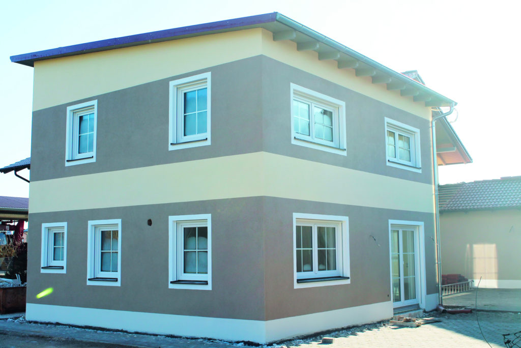 Zweistöckiges Haus mit Flachdach und beige- und cremefarbenen Außenwänden. Das Gebäude verfügt über mehrere weiß gerahmte Fenster und eine Glastür im Erdgeschoss. Es steht auf einer gepflasterten Fläche unter klarem Himmel.