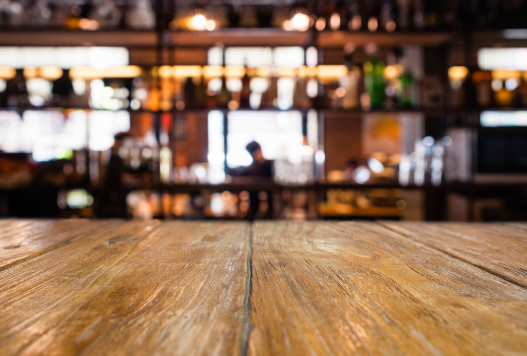 Eine Holzoberfläche im Vordergrund mit einem unscharfen Hintergrund einer schwach beleuchteten Bar. Regale sind mit verschiedenen Flaschen und Gläsern gefüllt und schaffen eine gemütliche, warme Atmosphäre.