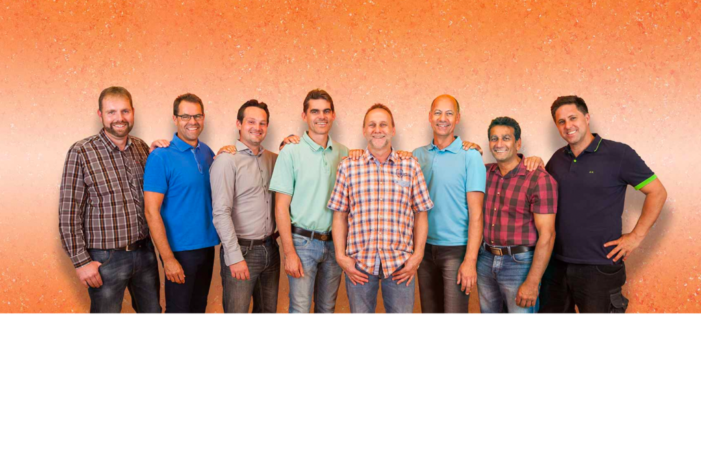 Acht Männer stehen Seite an Seite vor einem orange gesprenkelten Hintergrund, alle lächeln und tragen Freizeithemden in verschiedenen Farben wie Karomuster, Blau und Blaugrün.