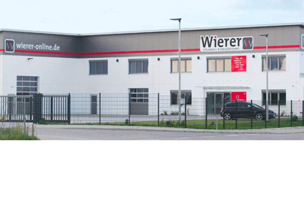 Ein modernes Industriegebäude mit den Schildern „Wierer“ und „wierer-online.de“ an den Wänden. Das Äußere zeichnet sich durch große Fenster, einen umzäunten Bereich und eine gepflasterte Auffahrt aus. Das Gebäude ist in Grau und Weiß mit roten Akzenten gehalten.
