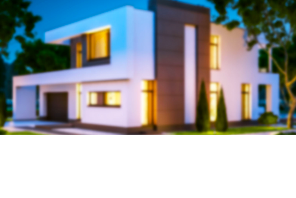 Unscharfes Bild eines modernen, zweistöckigen Hauses mit Flachdach und großen Fenstern vor einer Kulisse aus Bäumen und klarem Himmel. Das Haus wirkt durch das warme Licht gut beleuchtet und im Vorgarten gibt es etwas Vegetation.